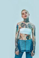 Tattoo and piercing. A white woman having piercing and tattoos looking into a camera while standing and wearing a denim overall with white top