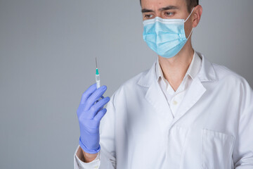 Doctor in white coat and blue mask holding coronavirus vaccine syringe in hand on gray background