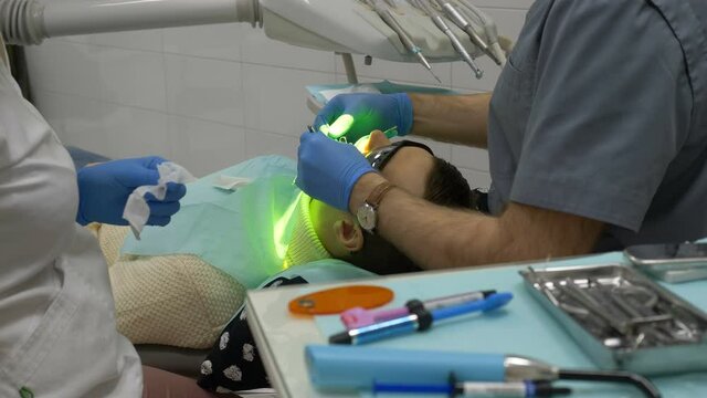 process of setting temporary seal, treatment at dentist office. young man in white sweater is lying in chair, doctor and assistant holding different devices in hands