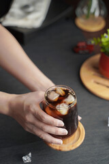 Hands holding a glass of espresso with lemon juice and fresh sliced lemon on wooden table and copy space, Summer Cocktail, Cold brew coffee or black tea. (close up, selective focus)