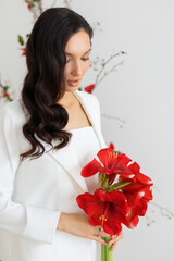 Bride in a white trouser suit posing against the background of an unusual composition of fresh flowers poinsetia for an chamber wedding ceremony. Hold bouquet of red amaryllis.
