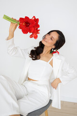 Fashionable bride in a white trouser suit and with a bouquet of red amaryllis sitting on chair....