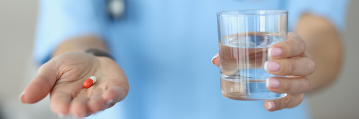 Woman doctor holding medicine capsule and glass of water in hands in clinic close-up. Regular medication intake concept