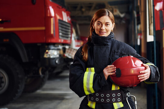 Holds red hat in hands. Female firefighter in protective uniform standing near truck