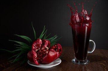 Red pomegranate juice splash in a beautiful glass, next to it lies a broken pomegranate on a wooden table with a dark background in the interior of the cafe. Drops juice scatter from the glass