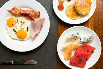 Delicious breakfast; sunny-side-up fried egg, ham, bacon, pancakes with fruits; on wooden table. Top view.