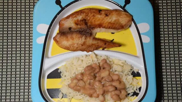 Tilapia fish fillet (Saint Peter) being placed on a children's plate with rice and beans.