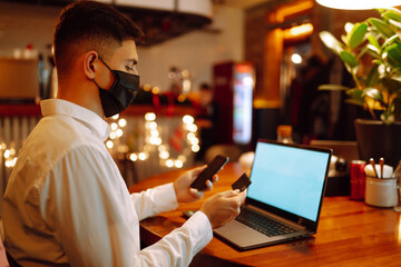 Young man in protective face mask using laptop and credit card for shopping online. Paying in internet during coronavirus quarantine. Digital payment. E-commerce store.