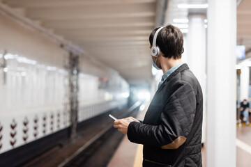 tattooed man in medical mask and wireless headphones holding smartphone in subway