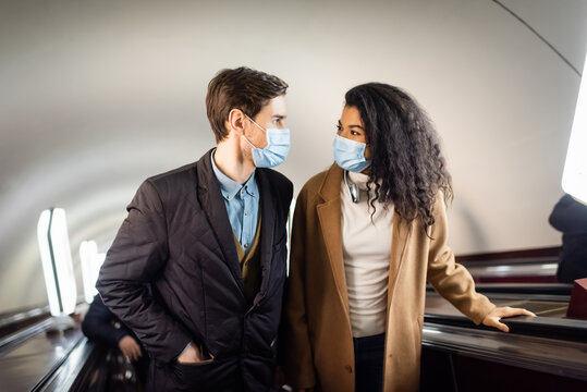 interracial couple in medical masks looking at each other on escalator in subway