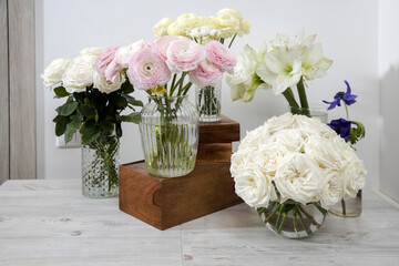 White roses, ranunculus, blue anemones, yellowish buttercups, lilies in round vases on the wooden boxes on the table for a special occasion as a kitchen decoration.