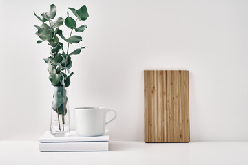 A mug on a stack of books and a transparent vase with eucalyptus branches, a wooden board against the wall. Eco-friendly materials in interior decor, minimalism. Copy space, mock up.