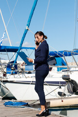 Female yachting dealer in marine blue suit holding notepad, standing outside a luxury sailboat...