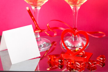 Legs of two glasses with champagne tied with red ribbons on a pink background next to a postcard.