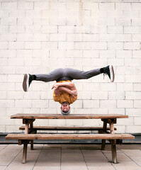 Caucasian white boy doing a headstand with yellow t-shirt head on top of a bench dancing break dance with gray brick background on the street