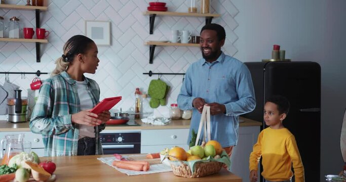 Young cheerful housewife cooking dinner when her family arriving home. Happy parents with son unpacking shopping bag with foods. Family in kitchen.