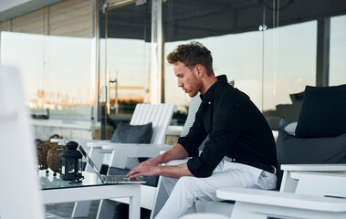 Sits by the table. Young man is outdoors at sunny daytime. Concept of vacation