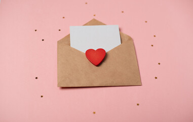 Craft envelope with a blank sheet of paper inside and red wooden heart on the rose background. Romantic love letter for the Valentine's day concept. Space for text.