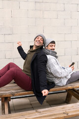 two white boys friends sitting on a bench listening to music with headphones and a smart phone on a bench enjoying and feeling the music dancing happy lifestyle