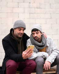 Two adult white caucasian friends wearing warm clothes and scarf listening to music with headphones looking at a yellow smart phone dancing laughing happy lifestyle sitting on a bench