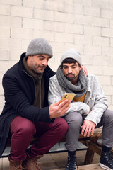 Two adult white caucasian friends wearing warm clothes and scarf listening to music with headphones looking at a yellow smart phone dancing laughing happy lifestyle sitting on a bench