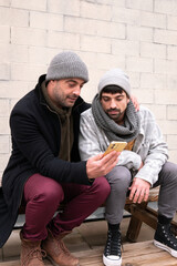 Two adult white caucasian friends wearing warm clothes and scarf listening to music with headphones looking at a yellow smart phone dancing laughing happy lifestyle sitting on a bench