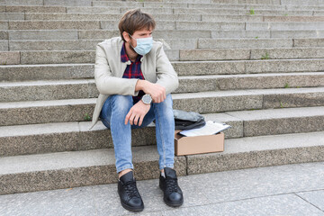 A man in a protective mask, fired from his job due to the economic crisis caused by the pandemic, sits on the steps of the street. Depression, unemployment - obrazy, fototapety, plakaty