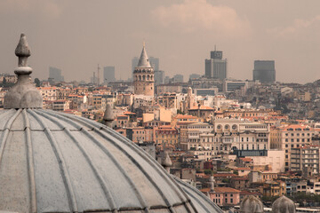Naklejka premium Orient cityscape. Sunlit old city centre. Houses with tiled roofs, a high round tower. A roof of the mosque out of focus in the foreground. Silhouettes of modern skyscrapers in the background