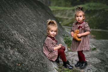 The friendship of two little girls. Girls are playing and having fun outdoors in a rocky area with teddy bears. Sisters in identical plaid dresses. The child's surprised eyes