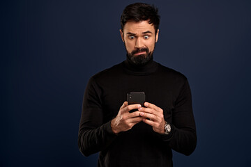 Concerned, worried young bearded man in sweater, biting lip nervously, holding smartphone...