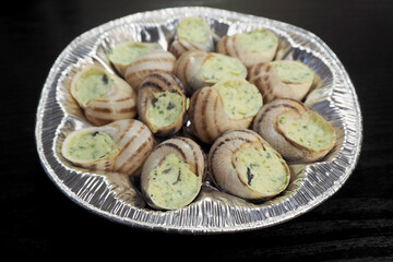 a lot of freshly prepared grape snails with green oil and garlic lie in a silver container on a black background of a side view .food in France snails in Burgundy
