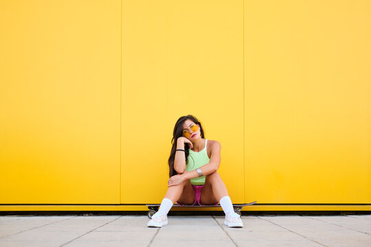 Portrait of beautiful girl sitting alone on longboard in front of yellow wall