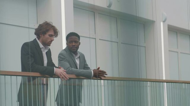 Medium shot of young African-American and Caucasian businessmen wearing elegant suits standing in business center hall leaning on glass railing and having chat