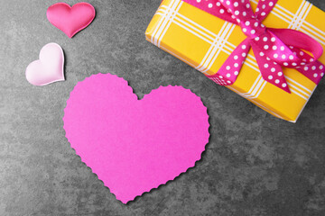 Blank heart shaped card and yellow gift box over grey stone background. Valentine’s day greeting