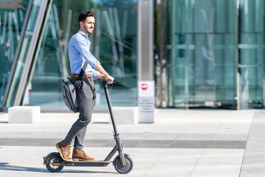 Young businessman with crossbody bag riding electric push scooter on footpath