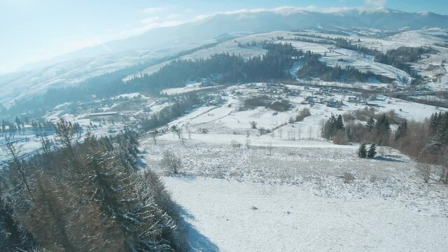 Aerial view of snow covered trees in the mountains in winter. Village at the foot of the mountain. Filmed on FPV drone