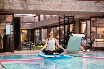 Female yoga teacher in lotus position on paddleboard over swimming pool