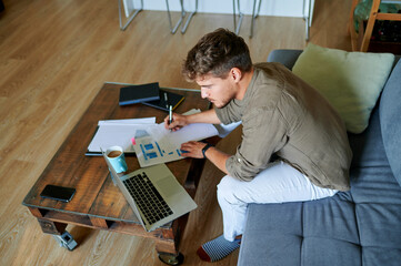 Male entrepreneur writing on paper while sitting with laptop at home