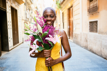 Happy woman in yellow dress holding flower bouquet while standing in city