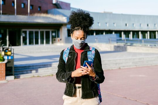 Student wearing protective face mask using smart phone while standing at city street