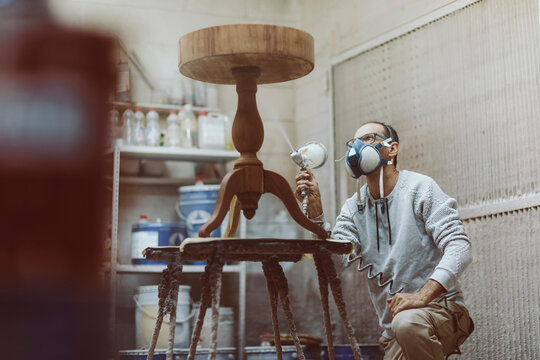 Carpenter with protective face mask lacquering table while working at workshop