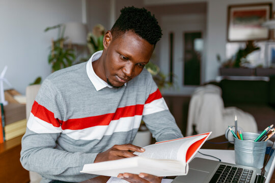Male entrepreneur reading book sitting by laptop at home