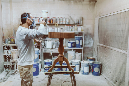 Manual worker wearing protective face mask spraying on table while standing at workshop