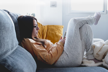 Young woman wearing glasses and yellow and white pyjamas typing on the smartphone, lying on the sofa. Indoors confinement, Covid 19