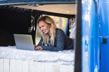 Smiling woman using laptop while lying on bed in van