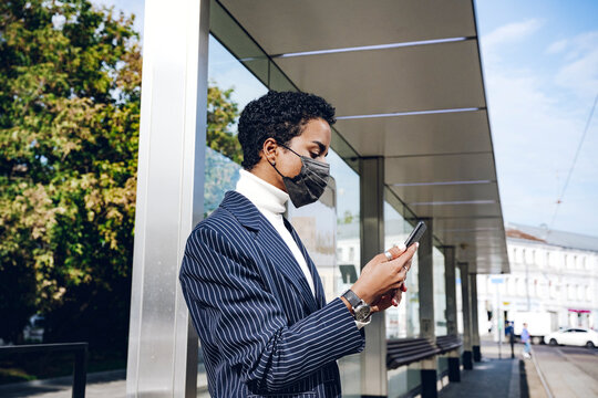Young businesswoman in protective face mask using mobile phone at bus stop