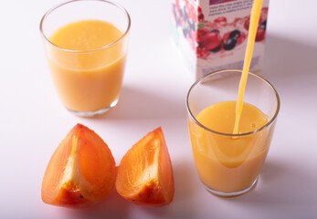 healthy persimon juice poured into glasses on white with two pieces of fruit at foreground and partially visible cartoon package.