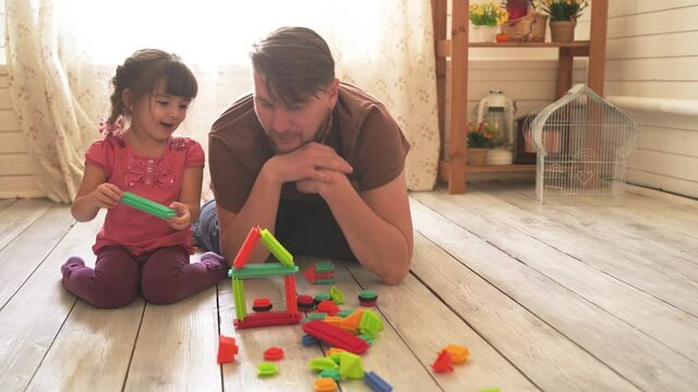 Little beautiful girl and her dad are sitting on the floor in the room and playing with a colorful constructor