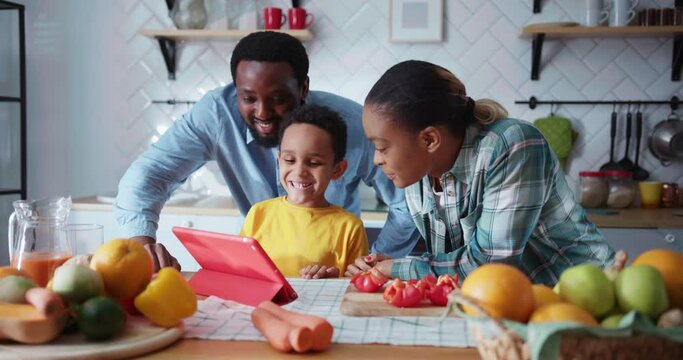 Joyful young family of parents and adorable son cooking in the kitchen talking to relatives online videochatting via tablet computer. Communication. Lockdown.