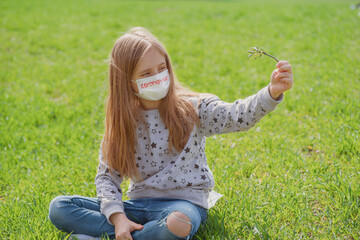 A little girl in a protective mask on her face is resting on the green grass in nature.Flu...